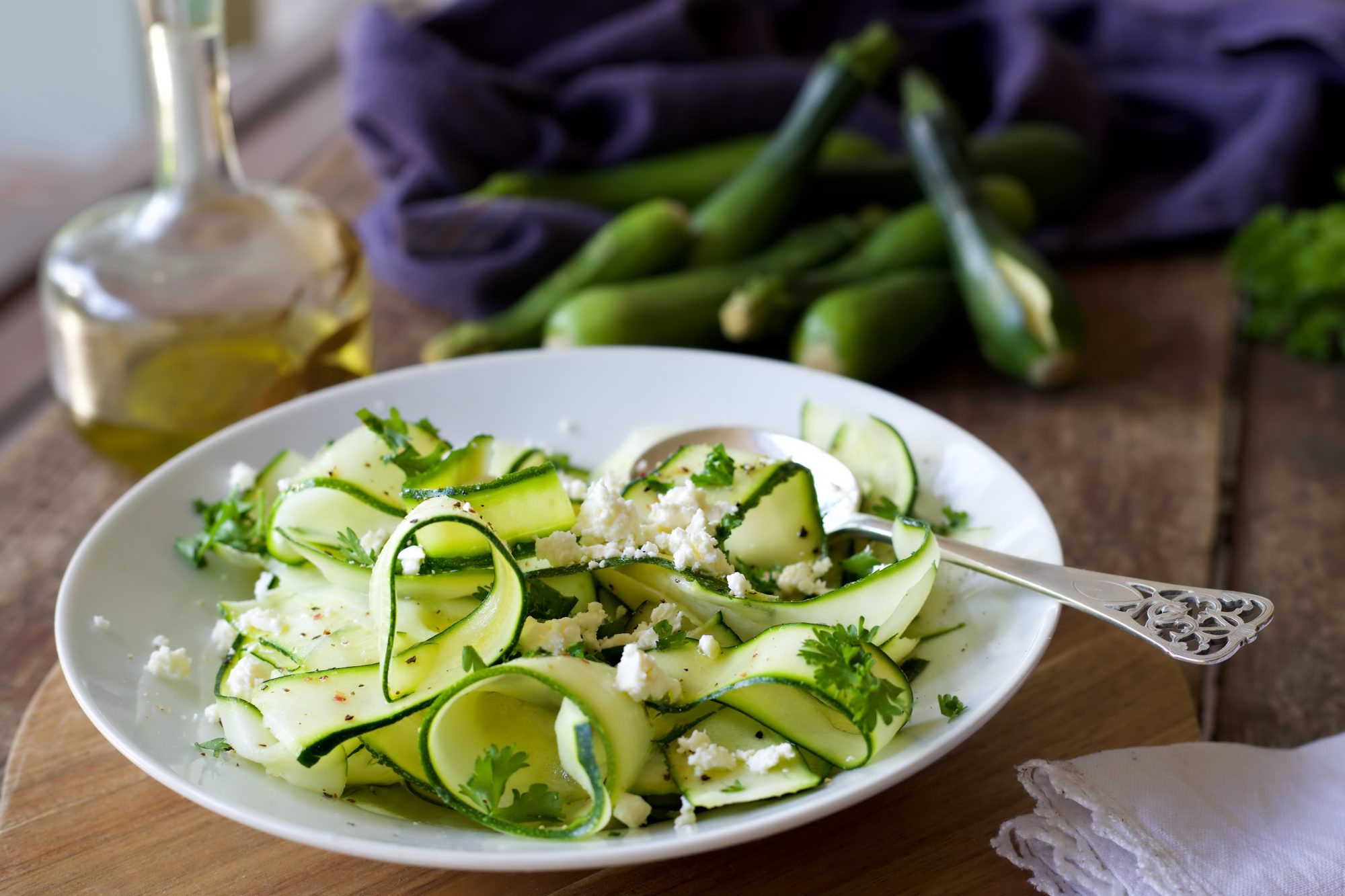 Zucchini ribbon salad in a white bowl with a silver spoon on a wooden table. Olive oil and whole courgettes on a blue cloth can be seen in the background